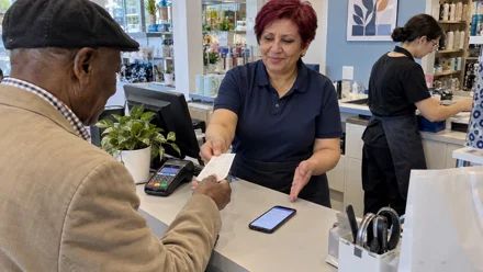 Alla cassa di un negozio, un cliente anziano con berretto riceve uno scontrino da una commessa in divisa scura, mentre sul bancone è appoggiato uno smartphone.