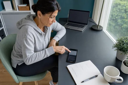 Donna seduta a una scrivania tocca lo schermo di uno smartphone, accanto un laptop, un quaderno e una tazza, luce naturale da finestra.