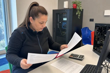 Donna con coda alta legge documenti seduta a una scrivania, accanto tastiera e calcolatrice, sullo sfondo un armadio server e una pianta.